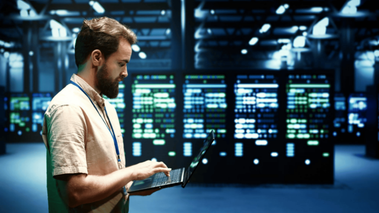 A bearded IT professional in a beige shirt and blue lanyard works on a laptop inside a modern data center. Behind him, rows of illuminated server racks glow with blue and green lights. The setting represents the infrastructure behind Cloud PBX systems, highlighting cloud-based communication technology in a secure, high-tech environment.